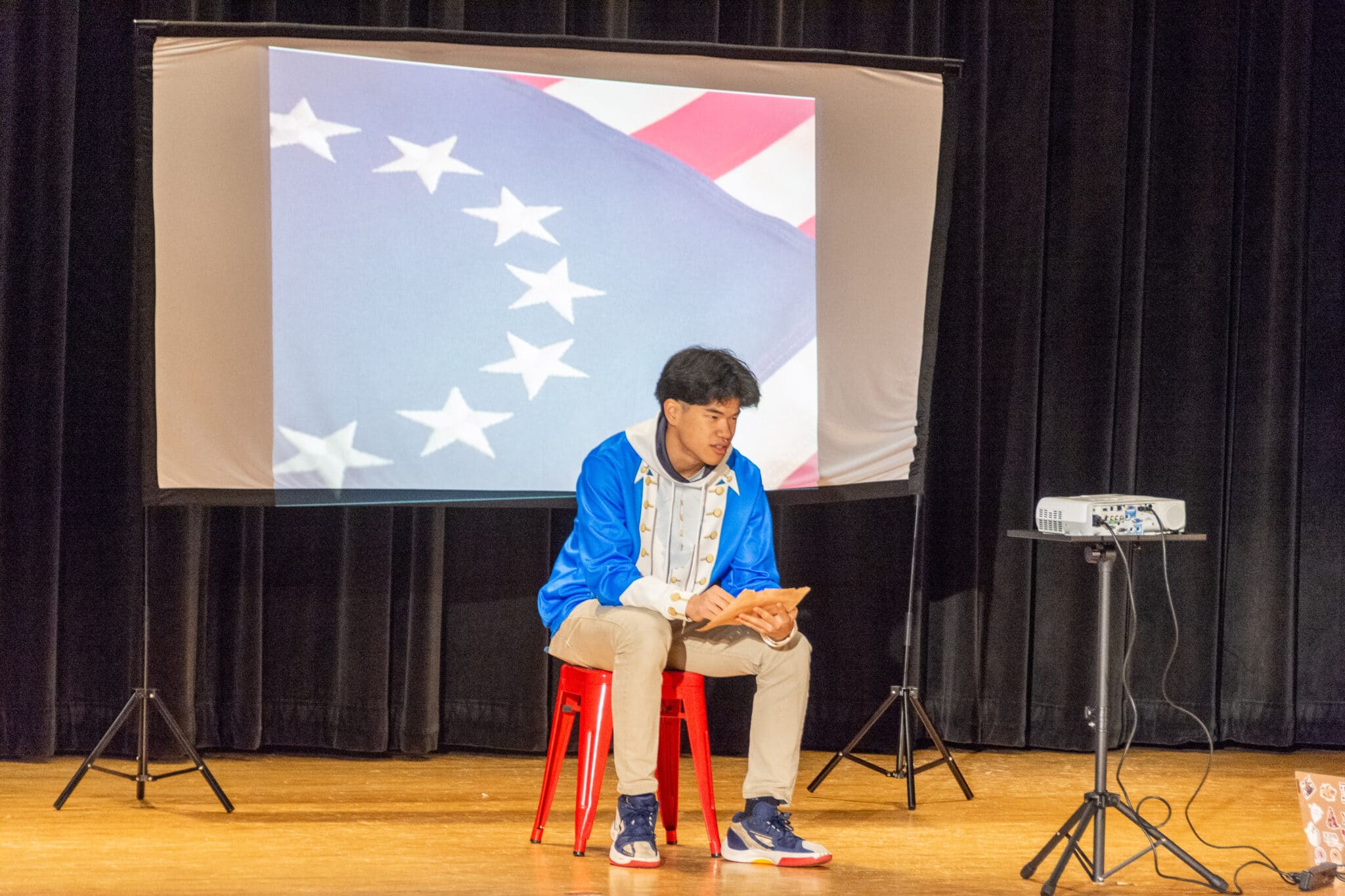 Student sitting on stage giving presentation behind PowerPoint screen with American flag on it